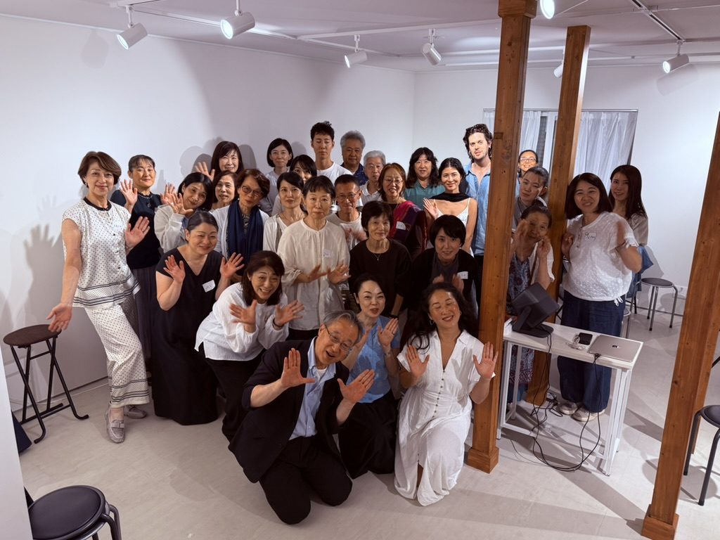 白い壁の会場で、参加者たちが両手を広げて笑顔で並んでいる集合写真。